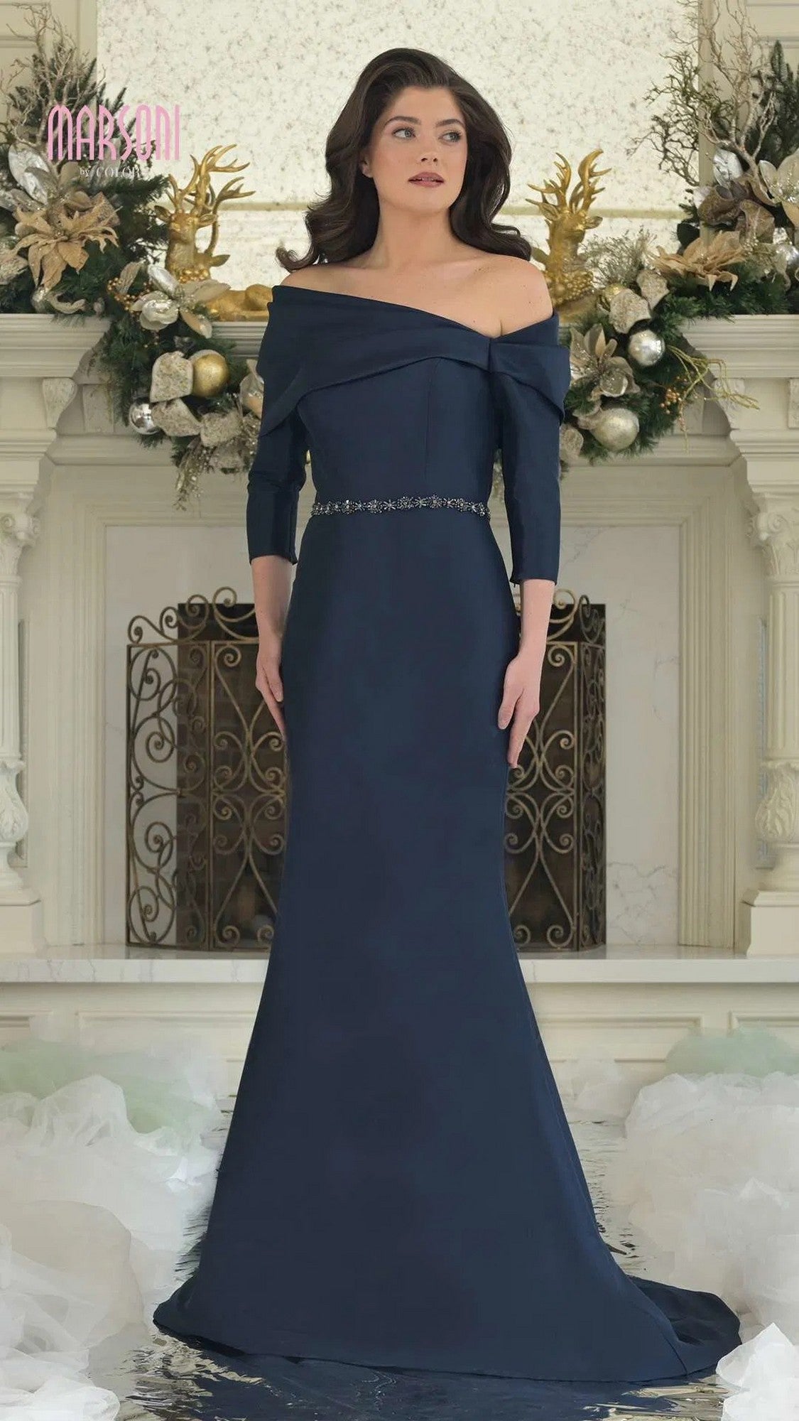 A woman in the Marsoni MV1375 long formal dress in navy, featuring an off-shoulder neckline and an embellished waist, stands before a mantel decorated with gold and white ornaments.